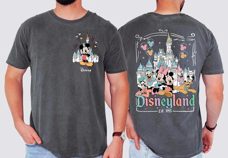 May include: A charcoal gray t-shirt featuring a small graphic of Mickey Mouse and a castle on the front chest with the word Daddy below it. The back displays a large graphic of Mickey, Minnie, Donald, Daisy, Goofy, and Pluto at Disneyland.