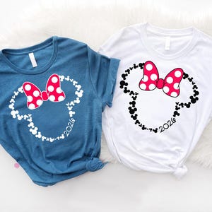 May include: A blue t-shirt and a white t-shirt, each featuring a graphic of a mouse head silhouette composed of smaller mouse icons. The graphic includes a red bow with white polka dots and the year 2026 printed in a script font at the bottom.