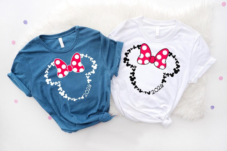 May include: A blue t-shirt and a white t-shirt, each featuring a graphic of a mouse head silhouette composed of smaller mouse icons. The graphic includes a red bow with white polka dots and the year 2026 printed in a script font at the bottom.