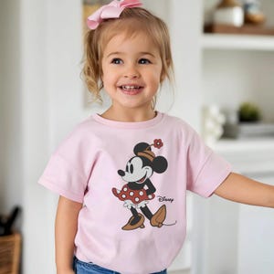 May include: A light pink short-sleeved t-shirt featuring a graphic of Minnie Mouse wearing a brown hat and brown shoes with a red polka-dot skirt. The character is depicted with a small red flower on her hat. The word Disney is printed in black.