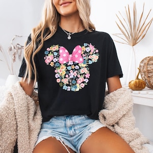 May include: A black short-sleeved t-shirt featuring a graphic of a mouse head silhouette composed of various colorful flowers. A large pink bow with white polka dots is positioned at the top center of the floral mouse head design.