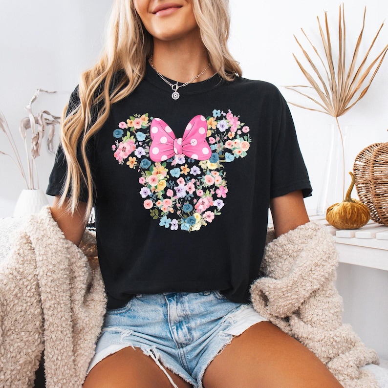 May include: A black short-sleeved t-shirt featuring a graphic of a mouse head silhouette composed of various colorful flowers. A large pink bow with white polka dots is positioned at the top center of the floral mouse head design.