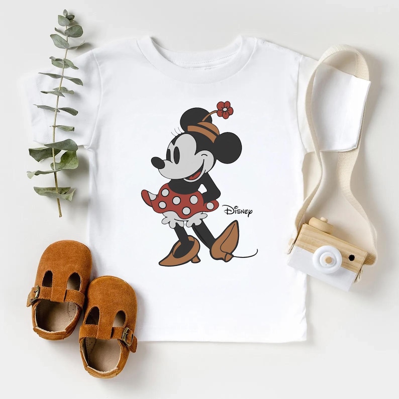 May include: A white short-sleeved t-shirt featuring a graphic of Minnie Mouse wearing a red polka-dot skirt, a brown hat with a red flower, and brown shoes. The word Disney is printed in black script next to the character on the front of the shirt.