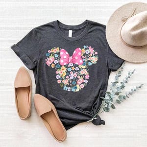 May include: A dark charcoal gray short-sleeve t-shirt featuring a central graphic of a mouse head silhouette composed of various colorful flowers, topped with a bright pink bow with white polka dots, accompanied by tan loafers and a tan hat.
