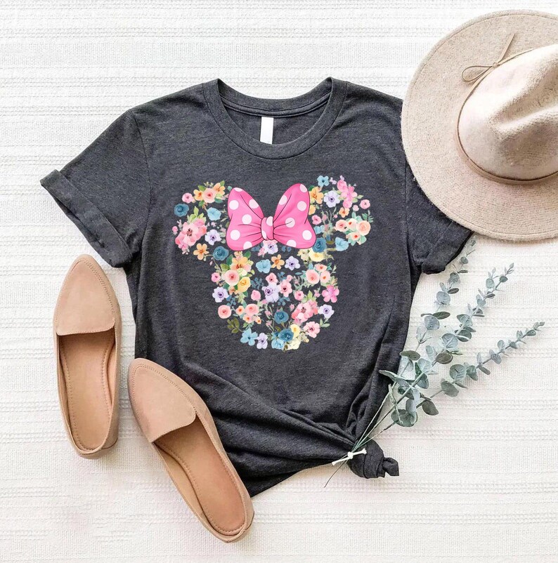 May include: A dark charcoal gray short-sleeve t-shirt featuring a central graphic of a mouse head silhouette composed of various colorful flowers, topped with a bright pink bow with white polka dots, accompanied by tan loafers and a tan hat.