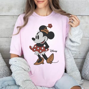 May include: A light pink t-shirt featuring a graphic of Minnie Mouse wearing a red polka dot skirt, brown shoes, and a hat with a flower. The character is depicted in a classic black and white style with the Disney logo printed in black text.