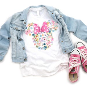 May include: A white short-sleeved t-shirt featuring a floral Minnie Mouse head silhouette with a pink polka-dot bow, layered under a light blue denim jacket, accompanied by a pair of pink canvas sneakers with white laces and rubber soles.