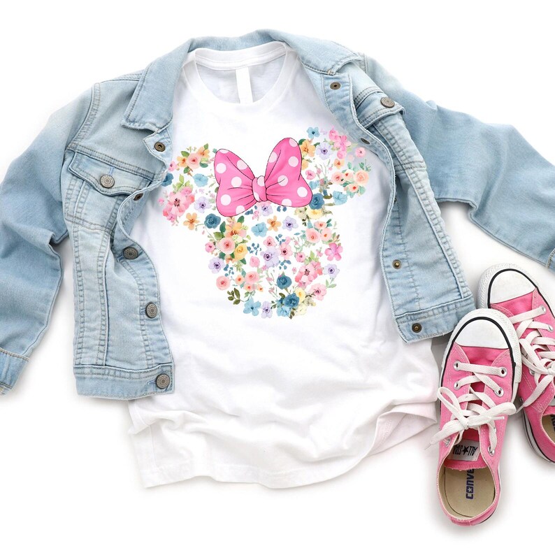 May include: A white short-sleeved t-shirt featuring a floral Minnie Mouse head silhouette with a pink polka-dot bow, layered under a light blue denim jacket, accompanied by a pair of pink canvas sneakers with white laces and rubber soles.