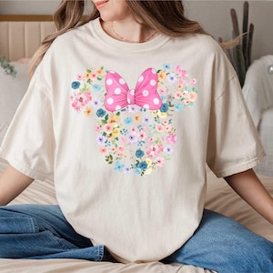 May include: A cream-colored short-sleeve t-shirt featuring a large graphic of a mouse head silhouette composed of various colorful flowers in shades of pink, blue, and yellow, topped with a bright pink bow with white polka dots.