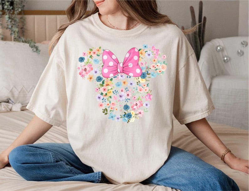 May include: A cream-colored short-sleeve t-shirt featuring a large graphic of a mouse head silhouette composed of various colorful flowers in shades of pink, blue, and yellow, topped with a bright pink bow with white polka dots.