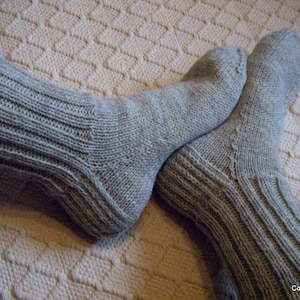 Könnte beinhalten: Ein Paar graue Stricksocken mit Rippenmuster. Die Socken sind umgeklappt und auf einer weißen, strukturierten Oberfläche abgelegt.
