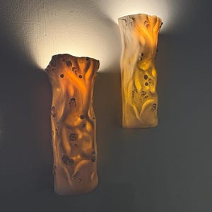 May include: Two wall sconces with a textured, organic design. The sconces are a warm amber color, with a mottled surface and irregular edges. They emit a soft, inviting glow, creating a cozy ambiance. The sconces are mounted on a dark gray wall.