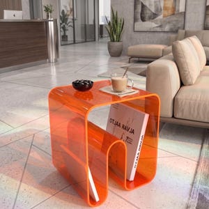 Könnte beinhalten: Ein durchscheinender orangefarbener Acryl-Beistelltisch mit geschwungenem U-förmigen Design. Der Tisch verfügt über ein integriertes Ablagefach, in dem ein Buch mit dem Titel OTJAA RAVJA steht. Eine kleine schwarze Schale und ein Glas mit beigem Getränk.