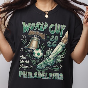 May include: Black t-shirt with a green and white graphic design for the World Cup 2026. The design includes a soccer ball, a soccer cleat, and the Liberty Bell. Text reads "World Cup 2026" and "the World plays in Philadelphia."