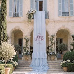 Può includere: Un lungo striscione in tessuto azzurro a balze che pende da un balcone. Il testo recita The HARRISON WEDDING in lettere rosso scuro, con un testo più piccolo sotto. Lo striscione si drappeggia lungo i gradini in pietra.