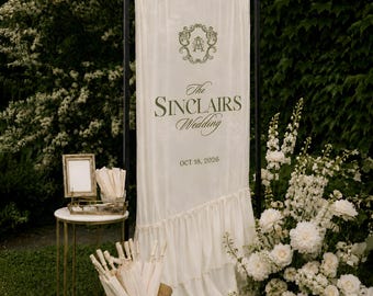 May include: A tall, off-white fabric wedding sign on a black metal frame featuring a decorative monogram, the text "The SINCLAIRS Wedding", and the date "OCT 18, 2026". The sign has a ruffled bottom edge and stands near a gold side table and bucket.