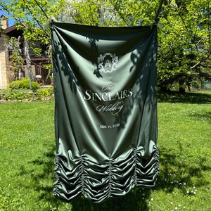 May include: A dark green wedding banner with the text "The Sinclairs Wedding" and the date "Aug 11, 2026". The banner is hanging outside on a sunny day, with a decorative bottom edge.