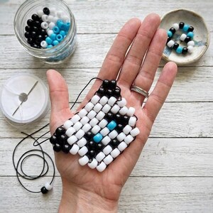 May include: A square-shaped beaded charm crafted from small white, black, and bright blue plastic pony beads arranged in a grid pattern. The charm is connected to a thin black cord, with additional loose beads in a glass jar and a small bowl.