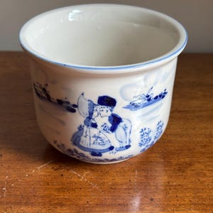 Peut inclure: Un pot ou bol en céramique blanche orné d'un motif peint à la main en bleu représentant deux personnages en tenue traditionnelle. La scène comprend des moulins à vent et des motifs floraux autour de l'extérieur. Le bord est souligné d'un fin liseré bleu.