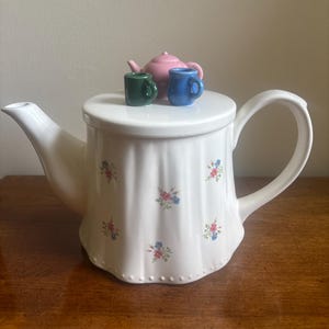May include: A white ceramic teapot with a fluted body and a delicate floral pattern featuring small pink and blue flowers. The lid is topped with three miniature decorative items: a small pink teapot, a dark green mug, and a light blue mug.