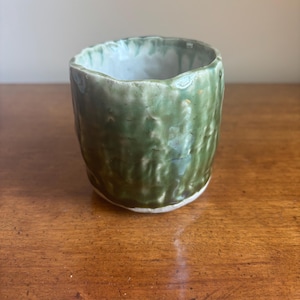 May include: A handmade ceramic cup with a textured, green exterior and a white interior. The cup has an irregular, organic shape and sits on a wooden surface. The glaze has variations in color, adding to its unique, artisanal appeal.