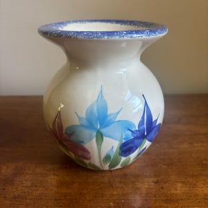 May include: A small, cream-coloured ceramic vase with a rounded body and a flared rim. The rim features a speckled blue pattern. The body is decorated with hand-painted floral motifs in shades of light blue, dark blue, and reddish-brown with green stems.