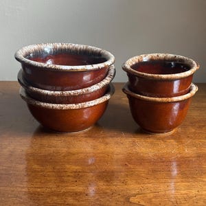 Pode incluir: Um conjunto de quatro tigelas de cerâmica com um acabamento castanho escuro brilhante e um esmalte de gotejamento de cor creme mais claro ao longo das bordas. As tigelas estão dispostas em duas pilhas, uma de duas tigelas maiores e outra de duas menores.