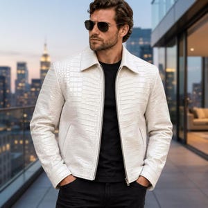 Può includere: Una giacca bianca con un motivo testurizzato in pelle di coccodrillo, dotata di colletto classico, maniche lunghe e chiusura frontale con zip color argento. Il capo è indossato sopra una t-shirt nera semplice e abbinato a pantaloni scuri.