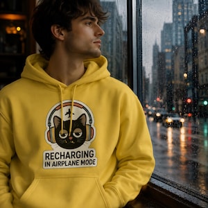 May include: A yellow hoodie with a graphic of a black cat wearing headphones and the text "RECHARGING IN AIRPLANE MODE." The hoodie has a front pocket and a drawstring hood. The person is looking out a window at a city street.