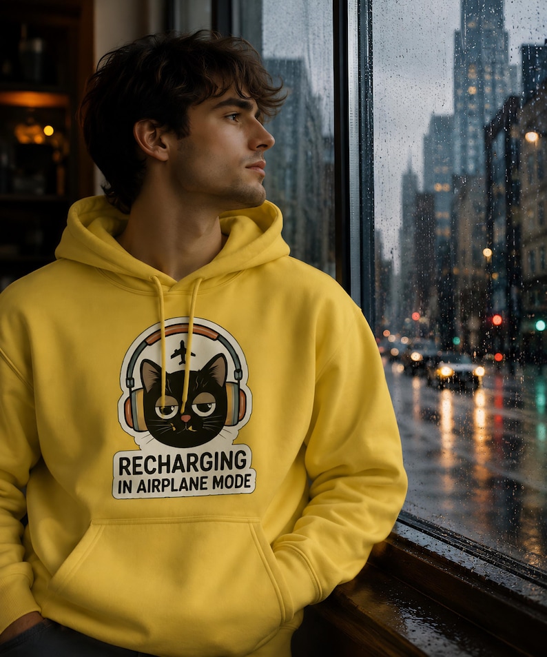 May include: A yellow hoodie with a graphic of a black cat wearing headphones and the text "RECHARGING IN AIRPLANE MODE." The hoodie has a front pocket and a drawstring hood. The person is looking out a window at a city street.