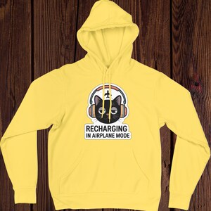 May include: A yellow hoodie with a front pocket and a hood. The graphic on the front features a black cat wearing headphones with a small airplane and the text "RECHARGING IN AIRPLANE MODE."
