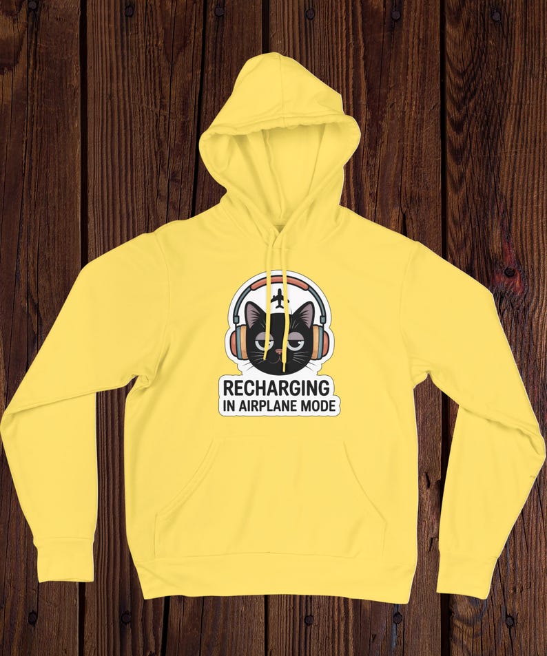 May include: A yellow hoodie with a front pocket and a hood. The graphic on the front features a black cat wearing headphones with a small airplane and the text "RECHARGING IN AIRPLANE MODE."