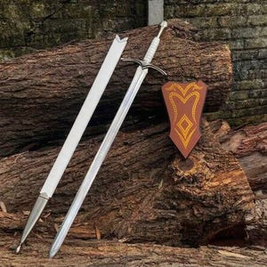 May include: A silver-toned metal sword with a white grip and a cross-shaped guard, accompanied by a matching white scabbard with silver metal fittings. Beside the sword is a brown, shield-shaped wall plaque featuring a yellow decorative pattern.
