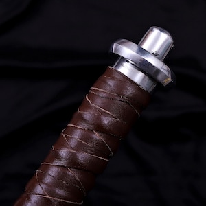 May include: Close-up of a sword handle. The handle is wrapped in brown leather with a crisscross pattern. The hilt is silver-colored metal with a circular guard and a rounded pommel.