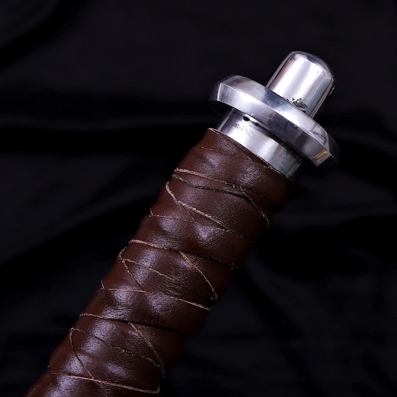 May include: Close-up of a sword handle. The handle is wrapped in brown leather with a crisscross pattern. The hilt is silver-colored metal with a circular guard and a rounded pommel.