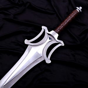 May include: A polished silver sword with a brown leather-wrapped handle. The blade is wide and has a distinctive shape with curved edges. The handle is wrapped in brown leather. The sword is displayed against a black background.