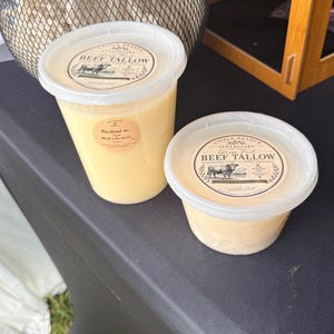 Puede incluir: Dos recipientes de plástico transparente llenos de sebo de res de color amarillo pálido. Cada recipiente tiene una etiqueta circular con el texto Little Branch Apothecary Grass Fed Beef Tallow y una ilustración de una vaca. El recipiente más grande tiene una pegatina adicional.