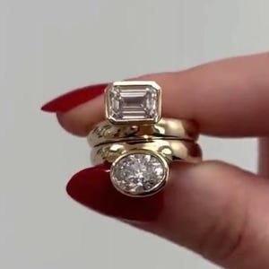 May include: A stack of two gold rings featuring bezel settings. The top ring displays a rectangular emerald-cut clear stone, while the bottom ring features a round brilliant-cut clear stone. Both bands are polished yellow gold with smooth surfaces.