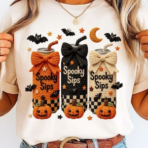Puede incluir: Una camiseta color crema con un gráfico de tres latas de bebida estilo ganchillo con la etiqueta Spooky Sips. Las latas son naranjas, negras y beige, decoradas con un lazo grande, una calabaza, murciélagos, estrellas y una luna creciente.