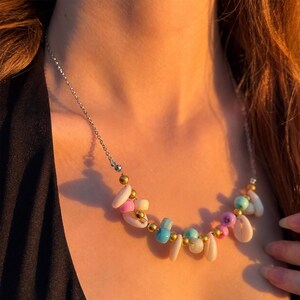 Peut inclure: Un collier composé d'une chaîne argentée ornée de petites perles de formes irrégulières dans des tons rose, bleu, violet et blanc, alternées avec de petites perles métalliques dorées et des éléments blancs rappelant des coquillages.