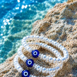 Peut inclure: Trois bracelets extensibles composés de perles nacrées blanches, d'entretoises argentées et d'une perle centrale en verre bleu foncé représentant un œil protecteur avec un iris blanc et bleu clair. Les bracelets sont empilés uniformément.