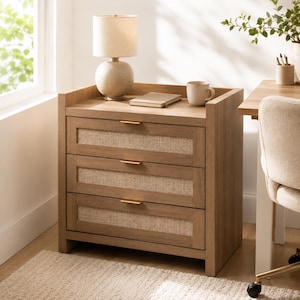 May include: A three-drawer wooden nightstand with woven cane inserts and brass-colored handles. A beige lamp, a small notebook, and a mug sit on top. The nightstand is in a well-lit room with a window.