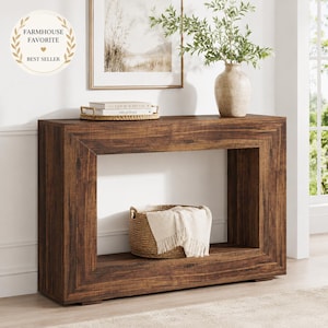 May include: A rectangular console table with a dark brown wood grain finish and a hollow center. The top surface holds a woven tray with two books, while a round woven basket containing a white fringed textile sits on the bottom shelf.