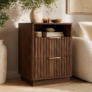 May include: A dark brown wooden nightstand with two drawers and a top shelf. The drawers have a vertical ribbed design and gold-colored handles. A vase with greenery, a candle, and decorative bowls sit on top.