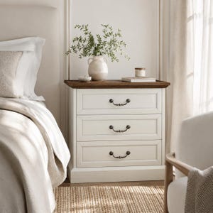 May include: A three-drawer nightstand with a white painted finish and a dark wood top. The drawers have ornate, dark metal handles. A white vase with greenery sits on top, along with a small candle and a book. The nightstand is next to a bed.