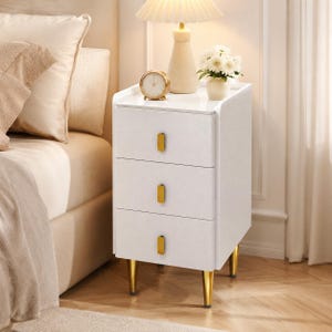 May include: A white nightstand with three drawers and gold-colored hardware and legs. A small gold clock, a lamp, and a vase of white flowers sit on top of the nightstand. The nightstand is next to a bed with beige bedding.
