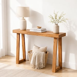 May include: A rectangular wooden console table with angled legs. On top sits a beige lamp with a textured base, two stacked books titled This is Home and Kinfolk, a small wooden bowl, and a vase with green stems. A woven basket with a beige throw blanket.