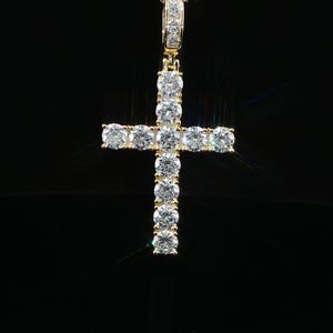 May include: A gold-toned cross pendant with clear, round-cut gemstones. The cross is suspended from a delicate chain, and the gemstones are set in a prong setting. The background is black.