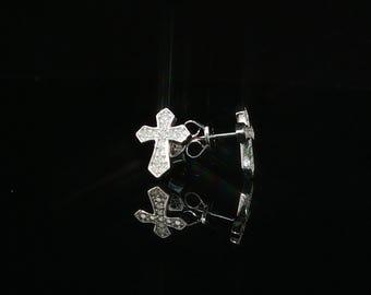 Può includere: Un paio di orecchini a lobo a forma di croce color argento con una superficie pavé composta da numerose piccole pietre chiare e scintillanti. Gli orecchini sono posizionati per mostrare il fronte e il perno, con un riflesso visibile sotto.