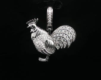 Può includere: Un ciondolo a forma di gallo color argento con finitura in metallo lucido, tempestato di numerose pietre chiare e scintillanti sul corpo e sulle piume della coda. Il pezzo include una contromaglia rettangolare anch'essa incastonata con piccole pietre.